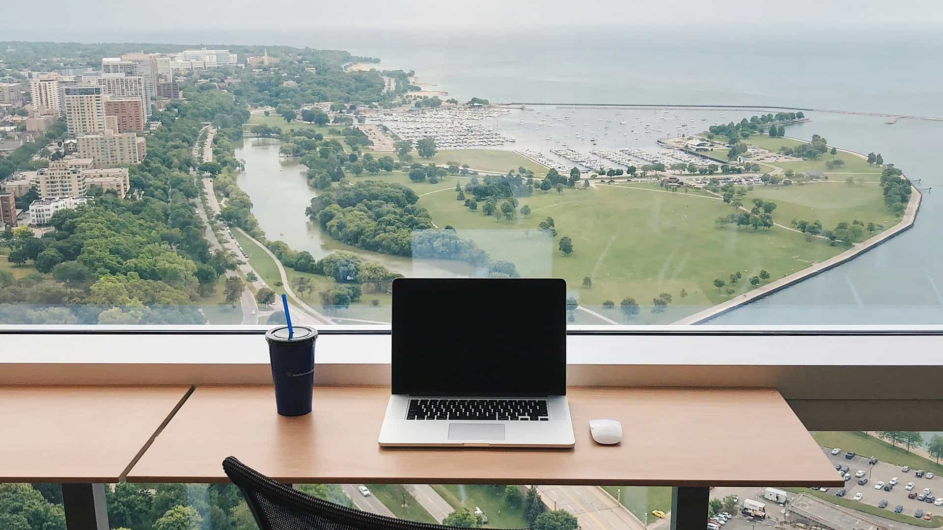 Ein Schreibtisch mit einem Laptop und einem Getränk darauf, der vor einem großen Fenster mit Blick über eine Küstenstadt steht.
